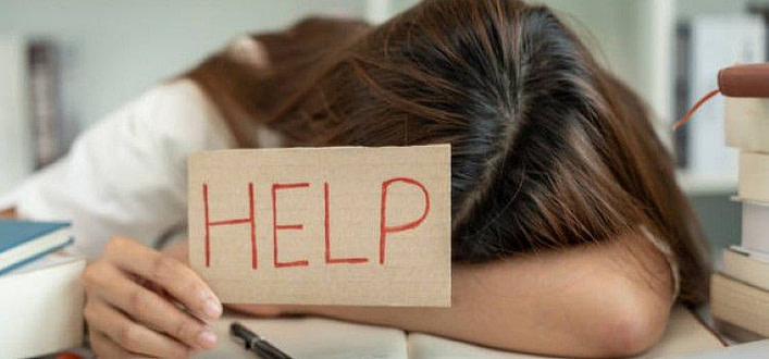 Giovane donna con testa accasciata sul braccio destro poggiato sul tavolo e cartello help in mano sinistra Giovane donna con testa accasciata sul braccio destro poggiato sul tavolo e cartello help in mano sinistra